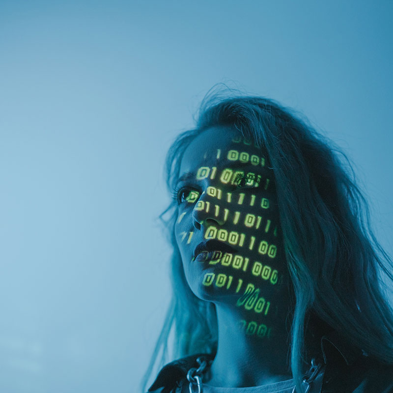 Artistic photo of a dimly lit woman looking into the distance. Computer code is brightly projected across her face.